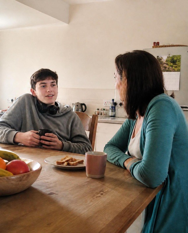 A teenage boy talking over a mug of tea at the kitchen table while his mother listens closely