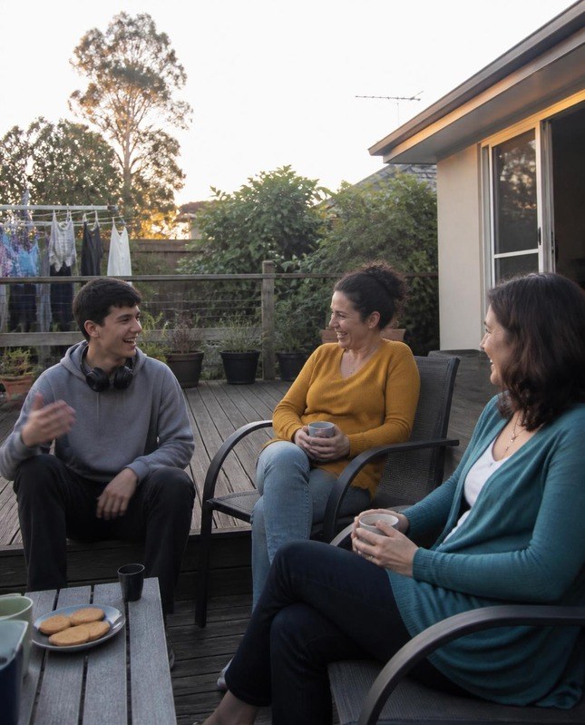 A teenage boy laughing and chatting with two women over tea and biscuits on a backyard deck at sunset