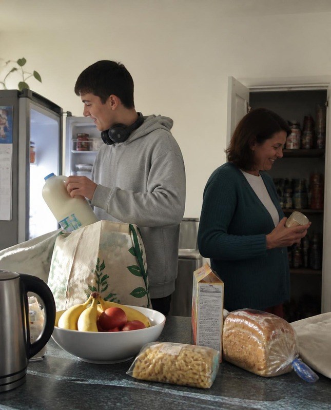A teenage boy and his mother unpacking groceries together in the kitchen, both smiling